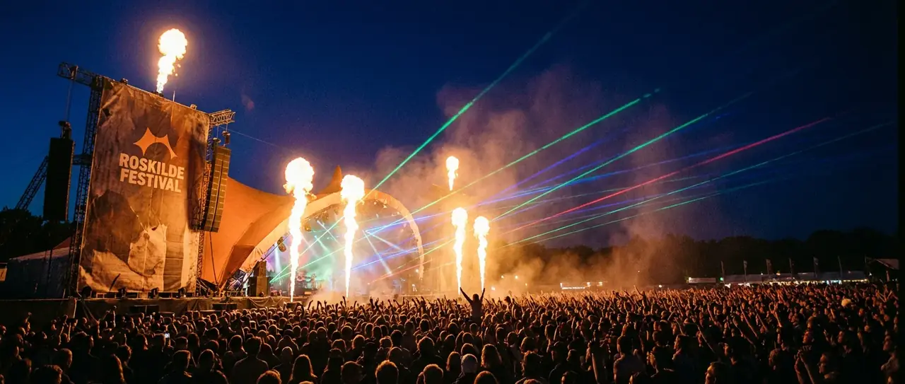 Publikum foran scenen på Roskilde Festival om aftenen, med flammer og laserlys over menneskemængden.
